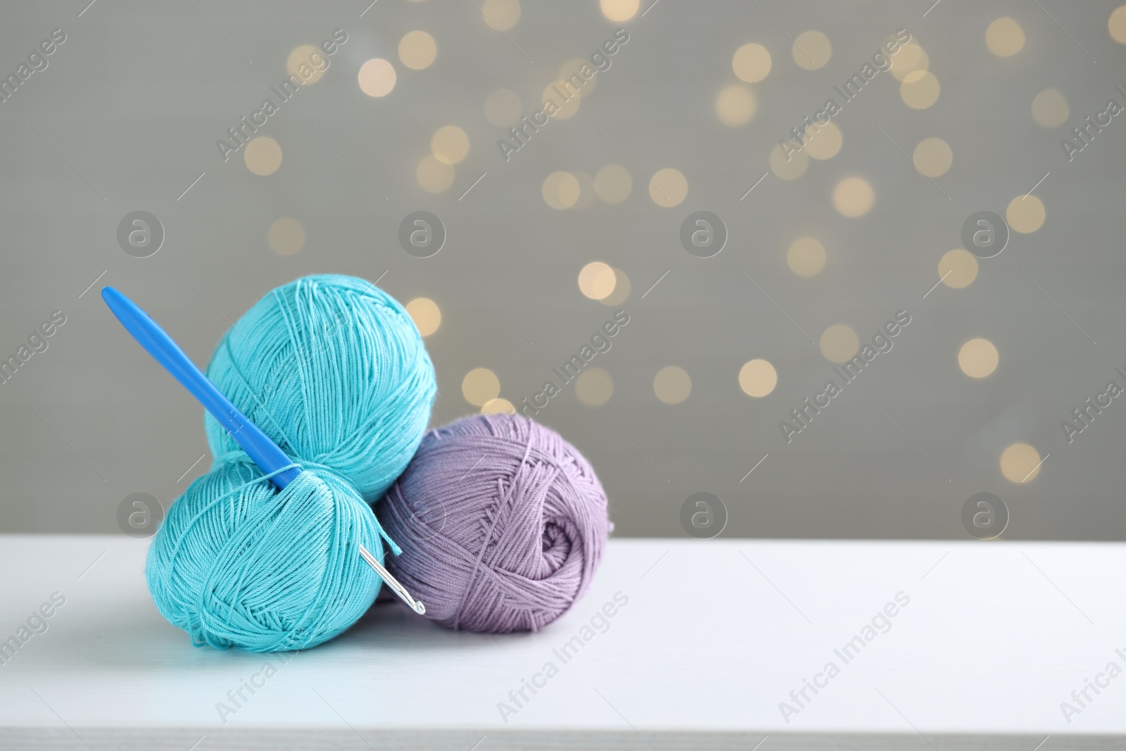 Photo of Colorful yarns and crochet hook on white table against grey background with blurred lights, space for text. Bokeh effect