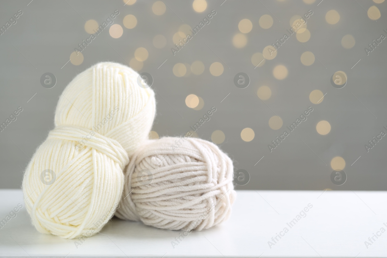 Photo of Skeins of yarn and knitting needles on white table against grey background with blurred lights, space for text. Bokeh effect