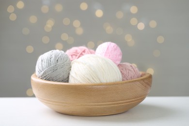 Photo of Balls of colorful yarn in bowl on white table against grey background with blurred lights, closeup. Bokeh effect