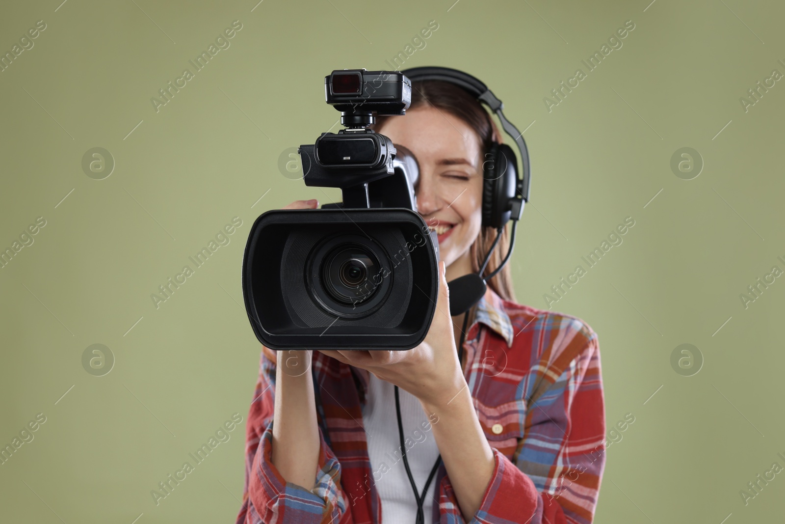 Photo of Happy woman with professional video camera and headset on pale olive background, selective focus