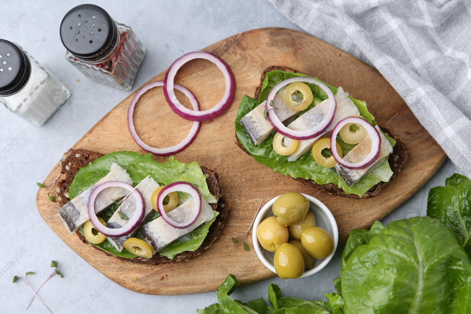 Photo of Tasty sandwiches with herring, onions, lettuce and olives on light grey table, flat lay