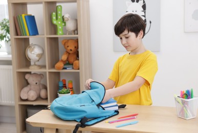 Photo of Boy packing backpack for school at wooden table indoors