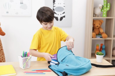 Photo of Boy packing backpack for school at wooden table indoors