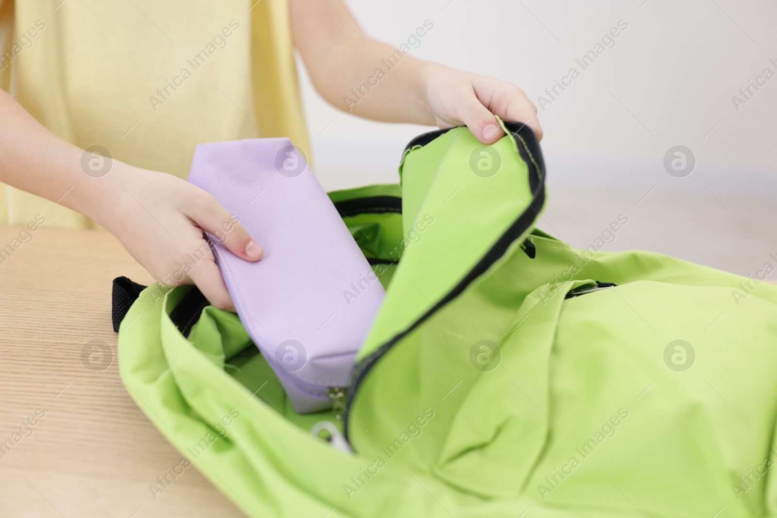 Photo of Little girl putting pencil case into backpack at wooden table indoors, closeup