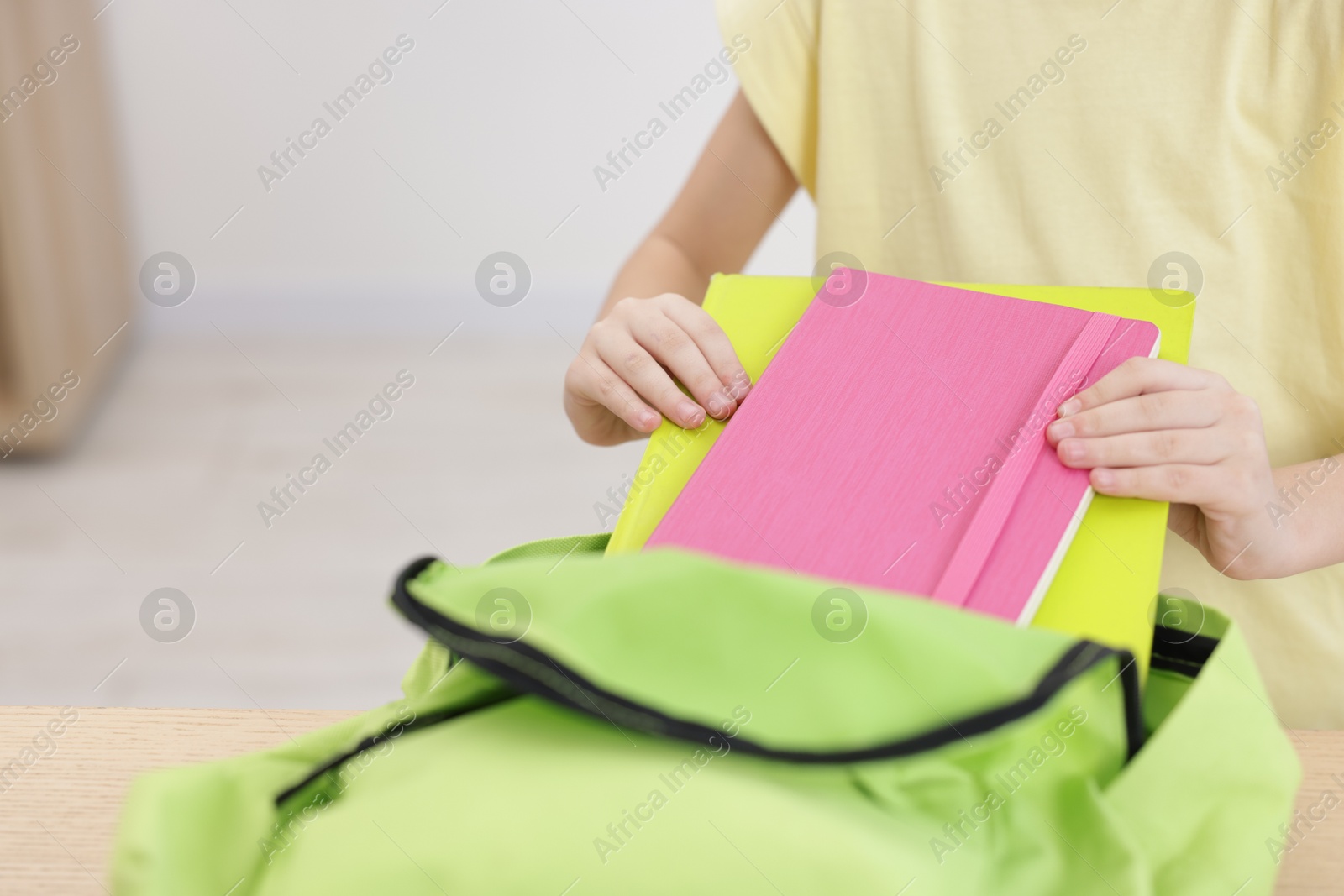 Photo of Little girl putting notebooks into backpack at wooden table indoors, closeup