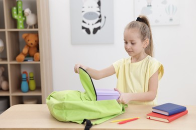 Photo of Little girl putting lunch box into backpack at wooden table indoors