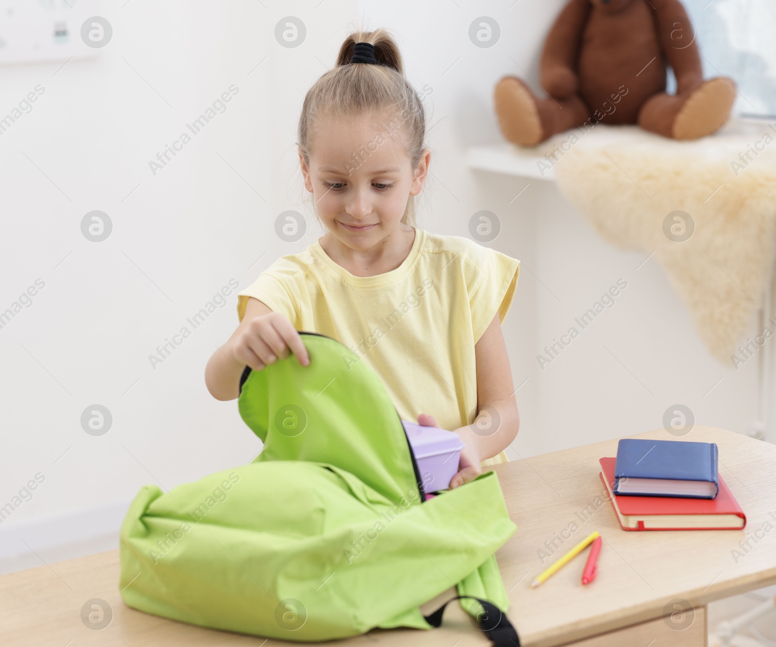 Photo of Little girl putting lunch box into backpack at wooden table indoors