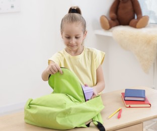 Little girl putting lunch box into backpack at wooden table indoors