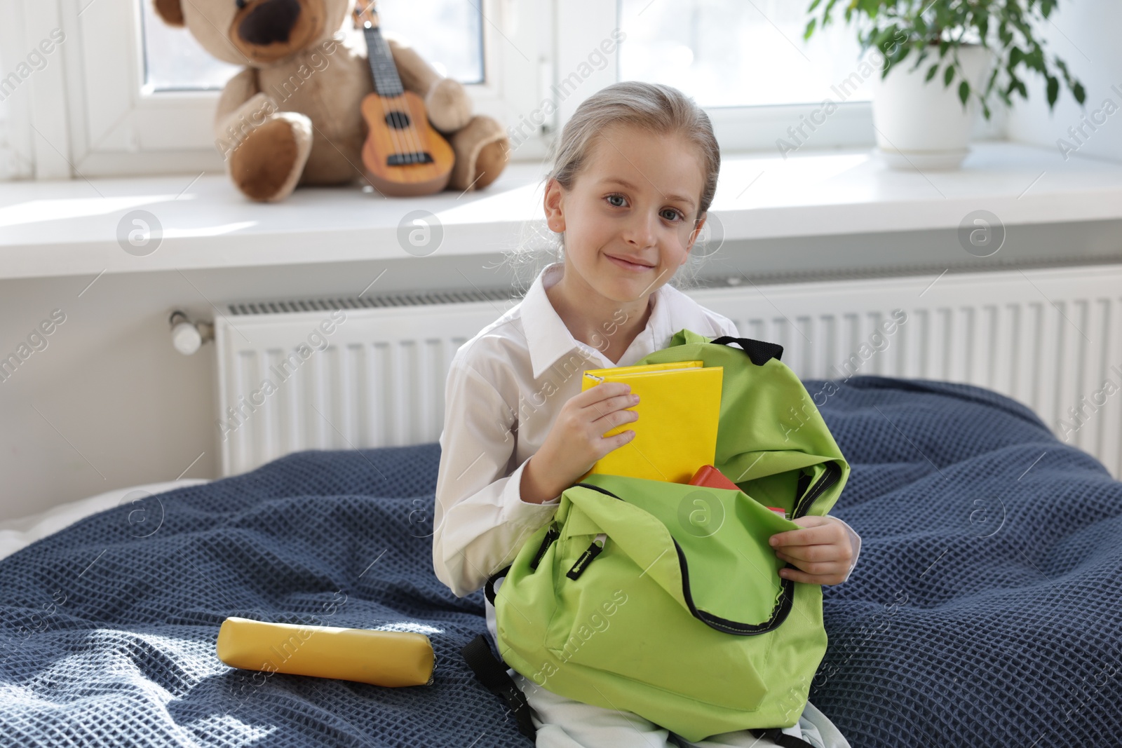 Photo of Girl packing book into backpack on bed indoors