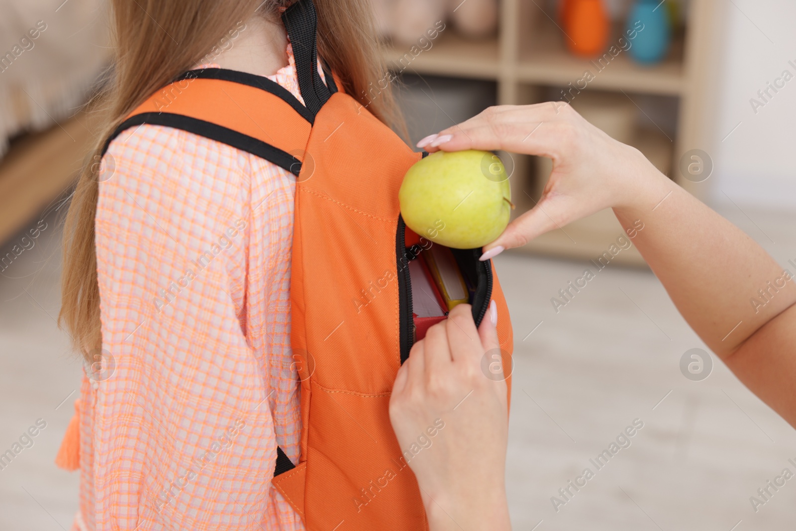 Photo of Mother putting apple into daughter’s backpack at home, closeup