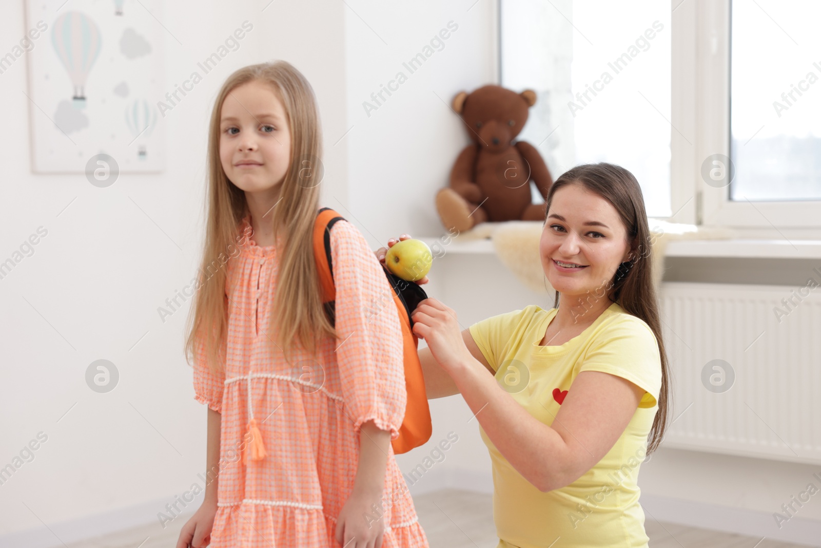 Photo of Mother putting apple into daughter’s backpack at home