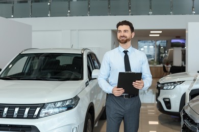 Photo of Happy salesman with clipboard near new cars in salon