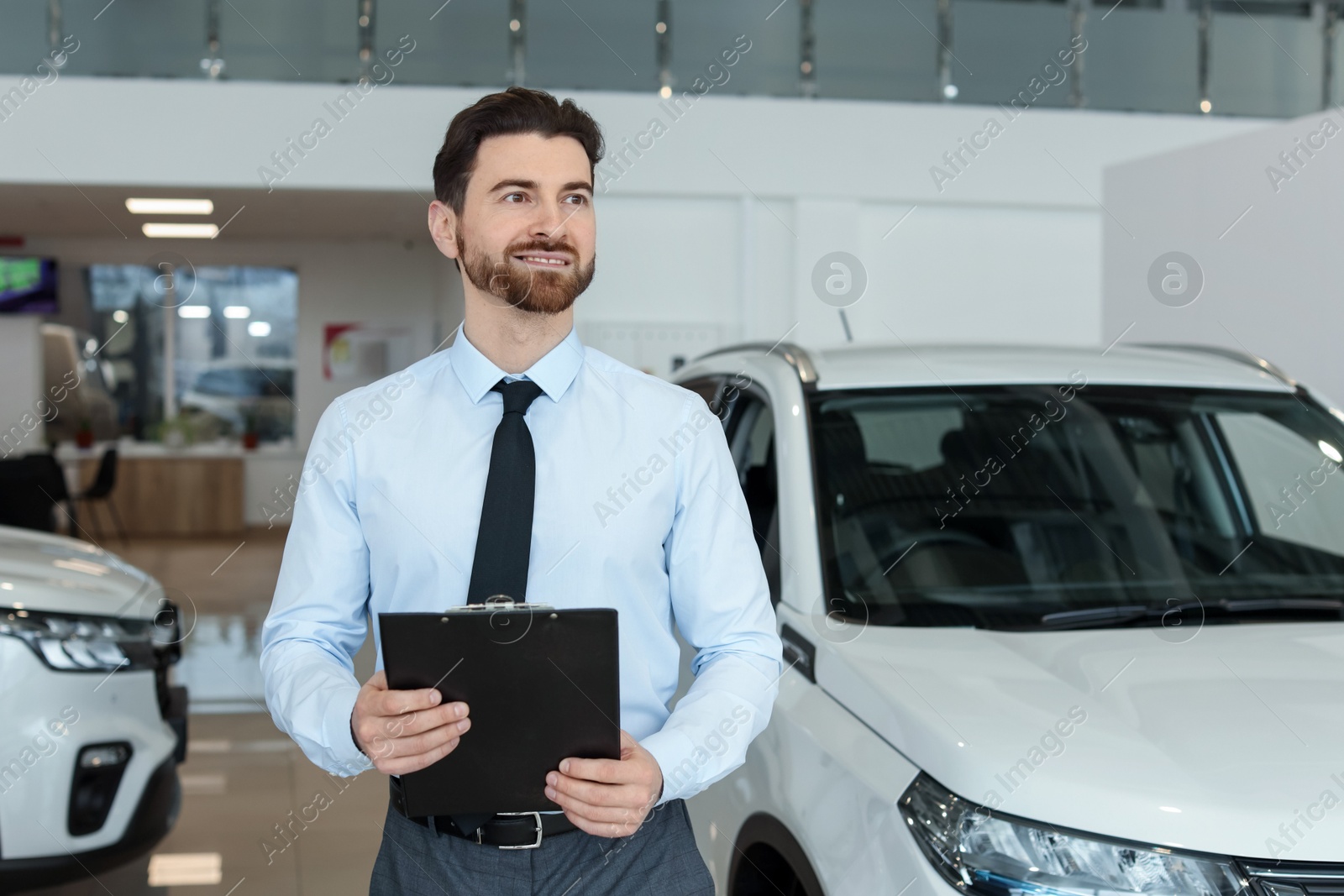 Photo of Happy salesman with clipboard near new cars in salon