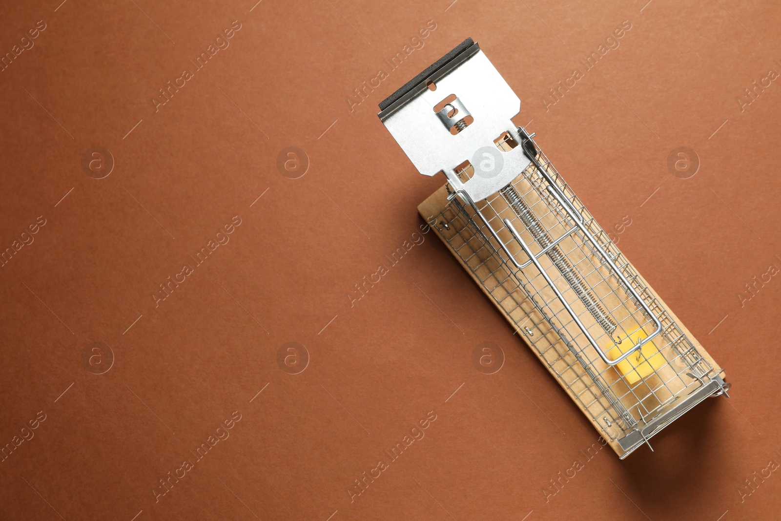 Photo of Metal mouse trap with piece of cheese on brown background, top view. Space for text