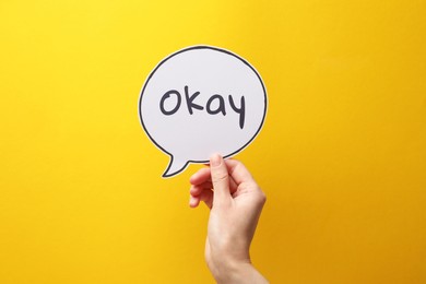 Image of Woman holding paper speech bubble with word Okay on yellow background, closeup