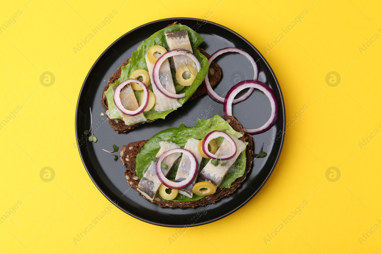 Photo of Tasty sandwiches with herring, onions, lettuce and olives on yellow background, top view