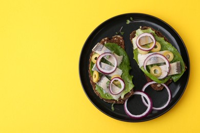 Photo of Tasty sandwiches with herring, onions, lettuce and olives on yellow background, top view. Space for text