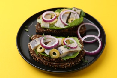 Tasty sandwiches with herring, onions, lettuce and olives on yellow background, closeup