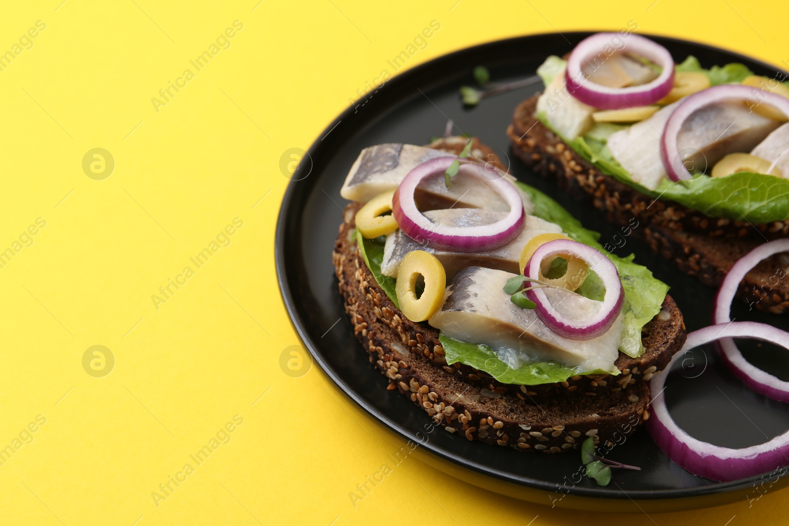 Photo of Tasty sandwiches with herring, onions, lettuce and olives on yellow background, closeup. Space for text