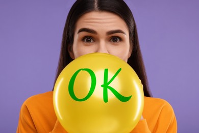 Image of Woman inflating yellow balloon with abbreviation OK on purple background