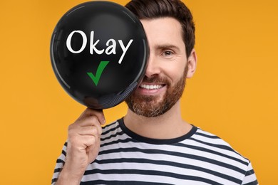 Image of Happy man holding black balloon with word Okay and check mark on yellow background