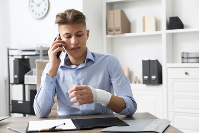 Photo of Man with wrist wrapped in medical bandage talking on smartphone at table indoors