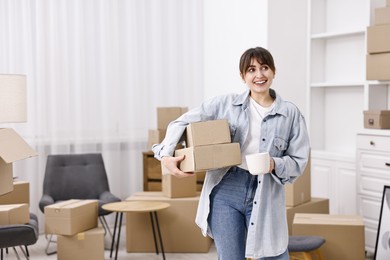 Photo of Moving day. Happy woman with cup of drink and cardboard boxes in her new home, space for text