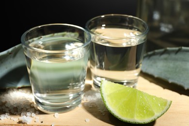 Photo of Tequila shots, slice of lime, salt and agave leaves on wooden table against black background, closeup