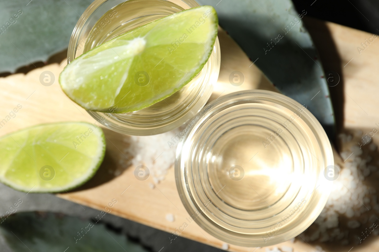 Photo of Tequila shots, slices of lime, salt and agave leaves on grey table, flat lay