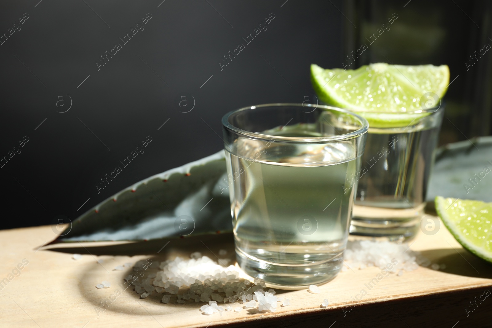 Photo of Tequila shots, slices of lime, salt and agave leaves on wooden table against black background, closeup. Space for text