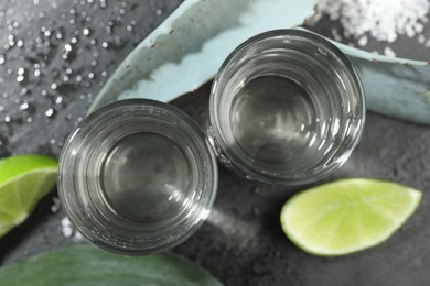 Photo of Tequila shots, slices of lime, salt and agave leaves on wet grey table, flat lay