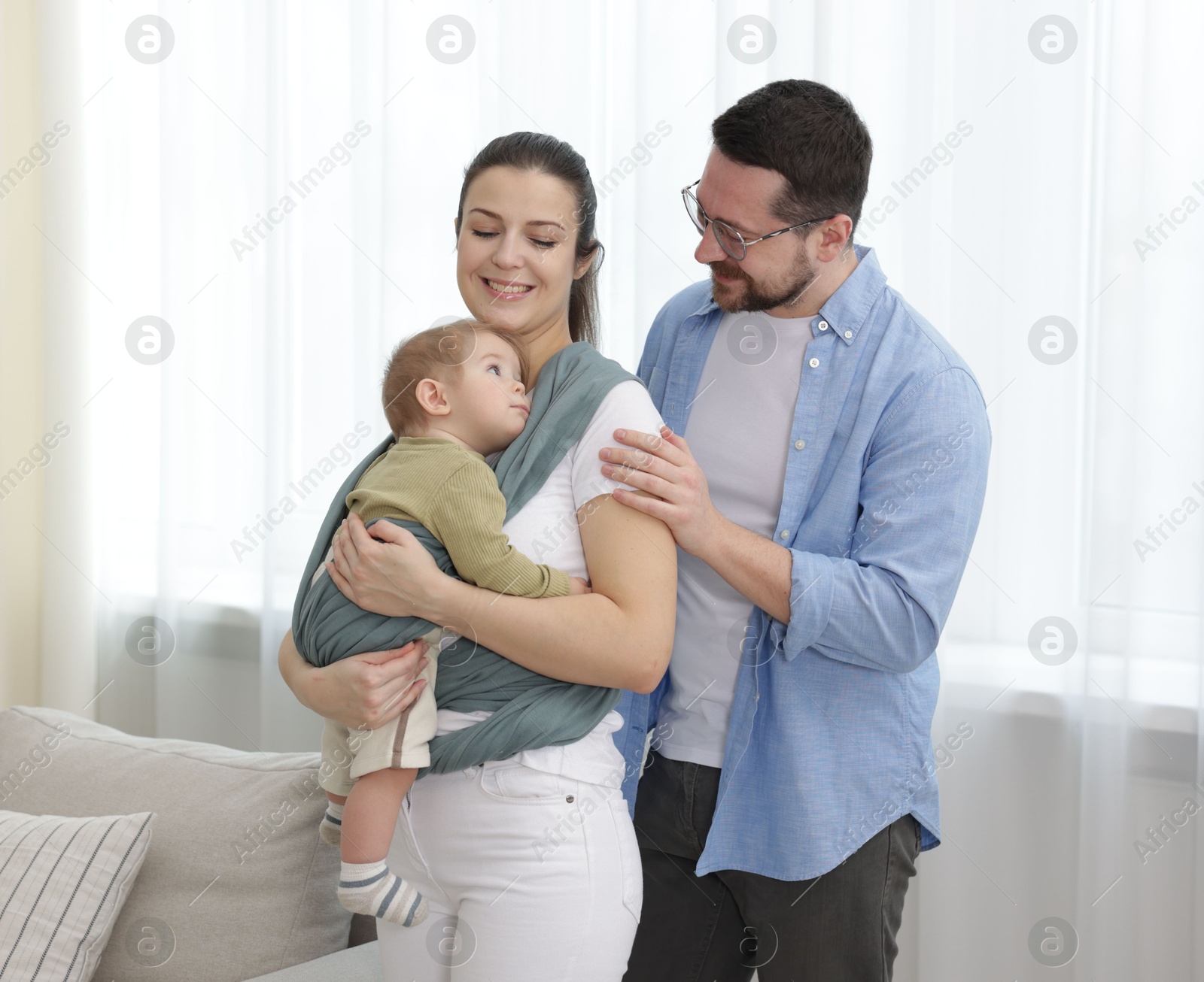 Photo of Portrait of happy family indoors. Mother holding her child in sling (baby carrier) at home