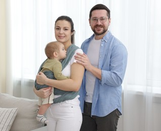 Photo of Portrait of happy family indoors. Mother holding her child in sling (baby carrier) at home