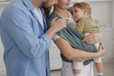 Parents with their baby indoors. Mother holding her child in sling at home, closeup