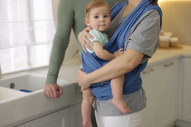 Parents with their baby indoors. Mother holding her child in sling at home, closeup