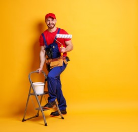 Photo of Professional painter with roller, bucket of paint and ladder on orange background. Space for text