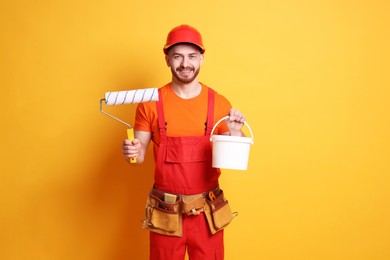 Professional painter with roller and bucket of paint on orange background