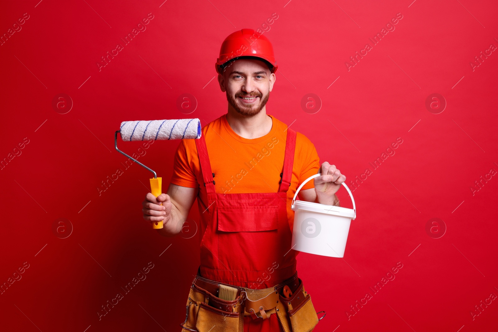 Photo of Professional painter with roller and bucket of paint on red background