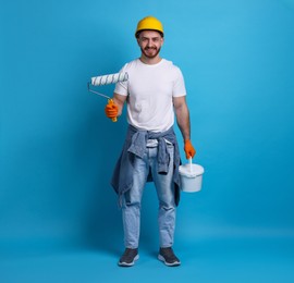 Photo of Man wearing hardhat with roller and bucket of paint on blue background