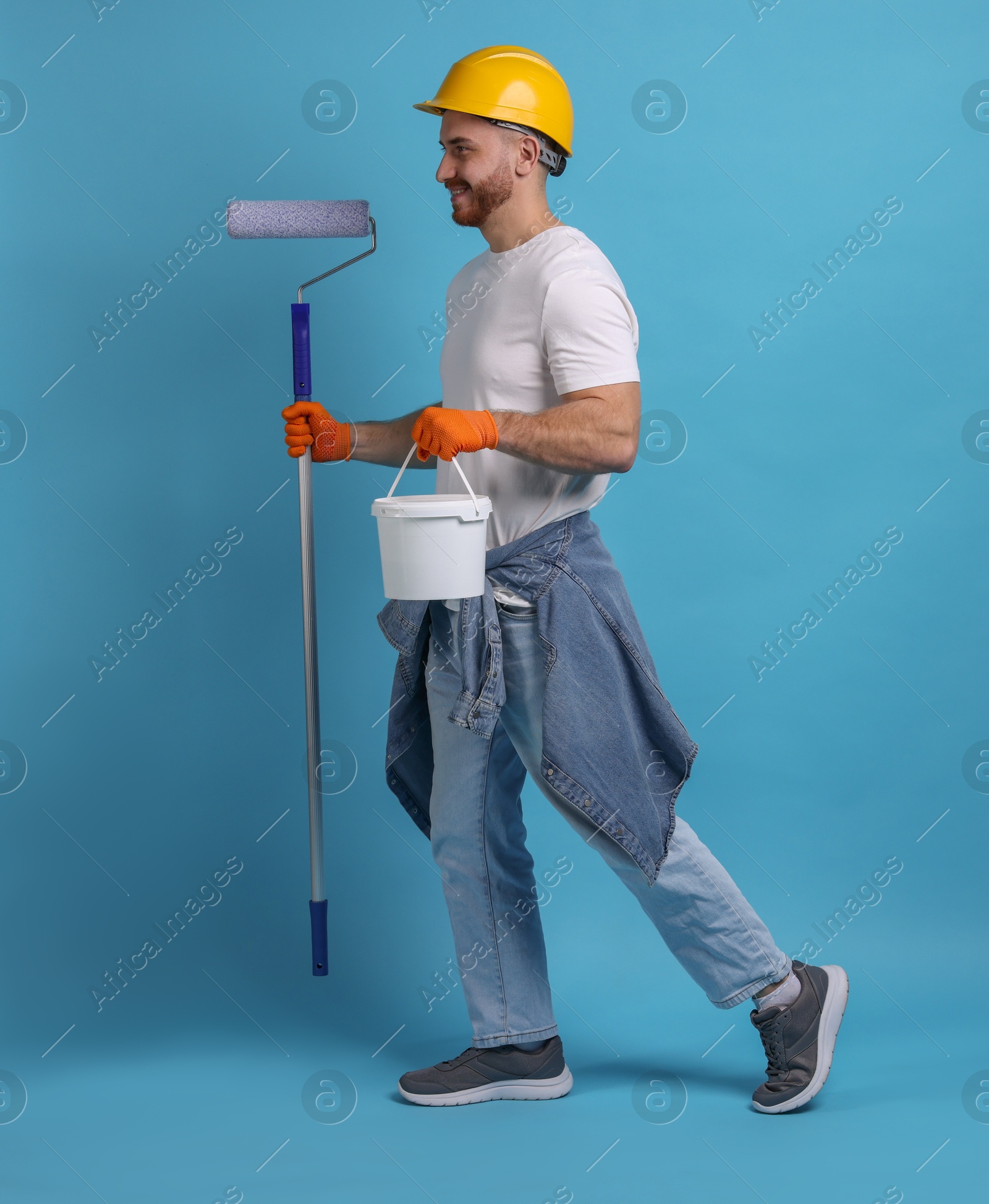 Photo of Man wearing hardhat with roller and bucket of paint on blue background