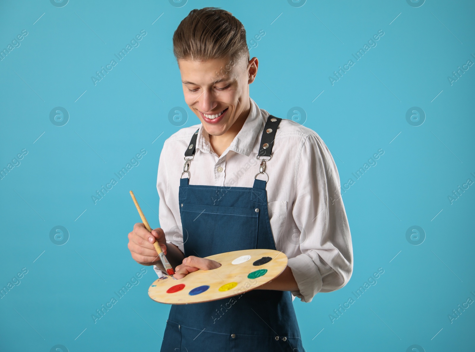 Photo of Smiling man with wooden palette and paintbrush on light blue background