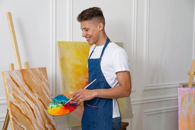 Smiling man with palette and paintbrush drawing picture indoors