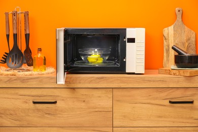 Photo of Open microwave oven with bowl of water and lemons on wooden cabinet near orange wall indoors