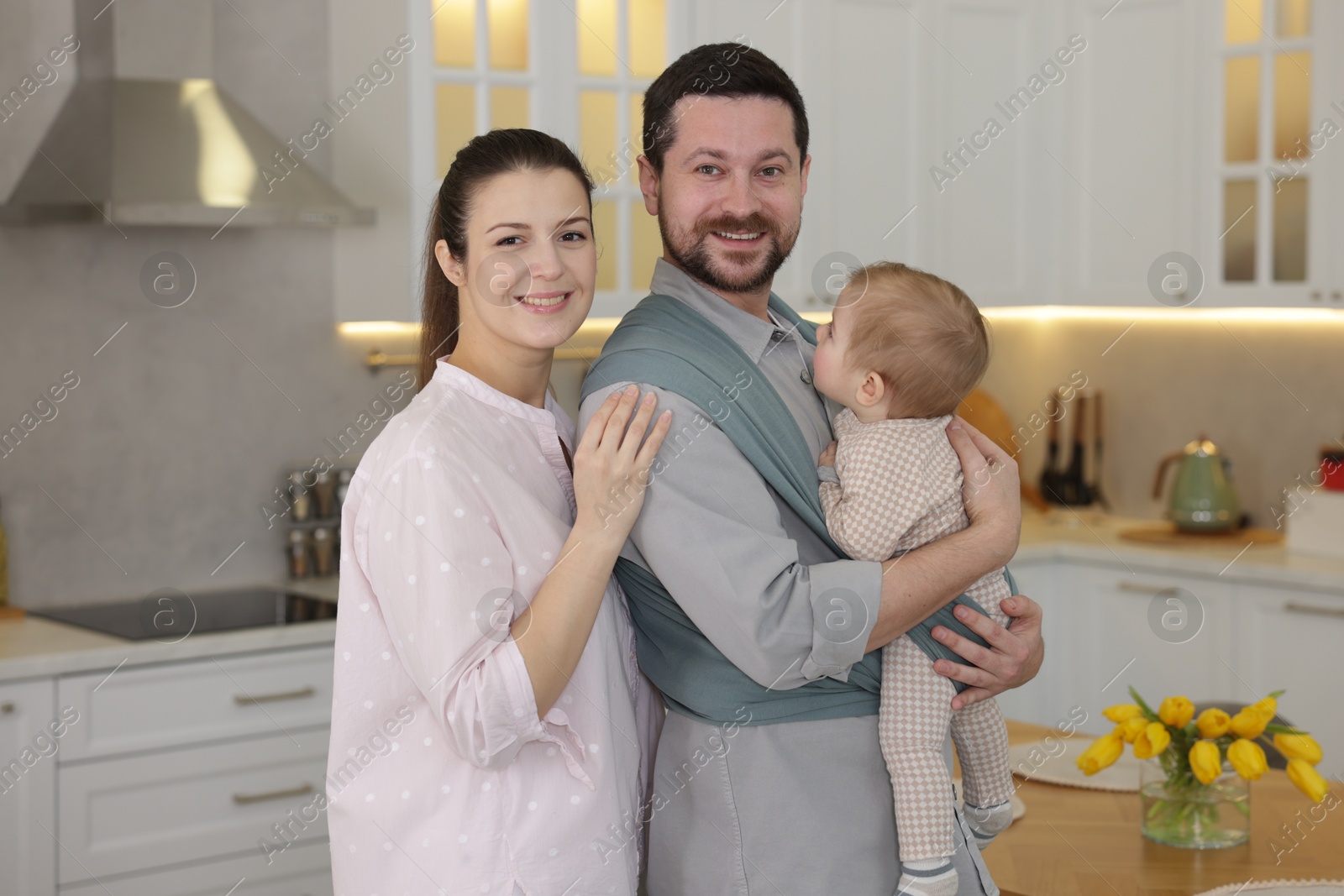 Photo of Portrait of happy family indoors. Father holding his child in sling (baby carrier) at home