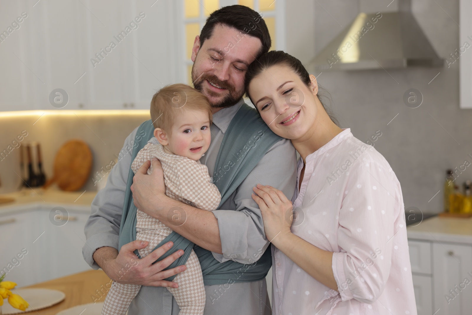 Photo of Portrait of happy family indoors. Father holding his child in sling (baby carrier) at home