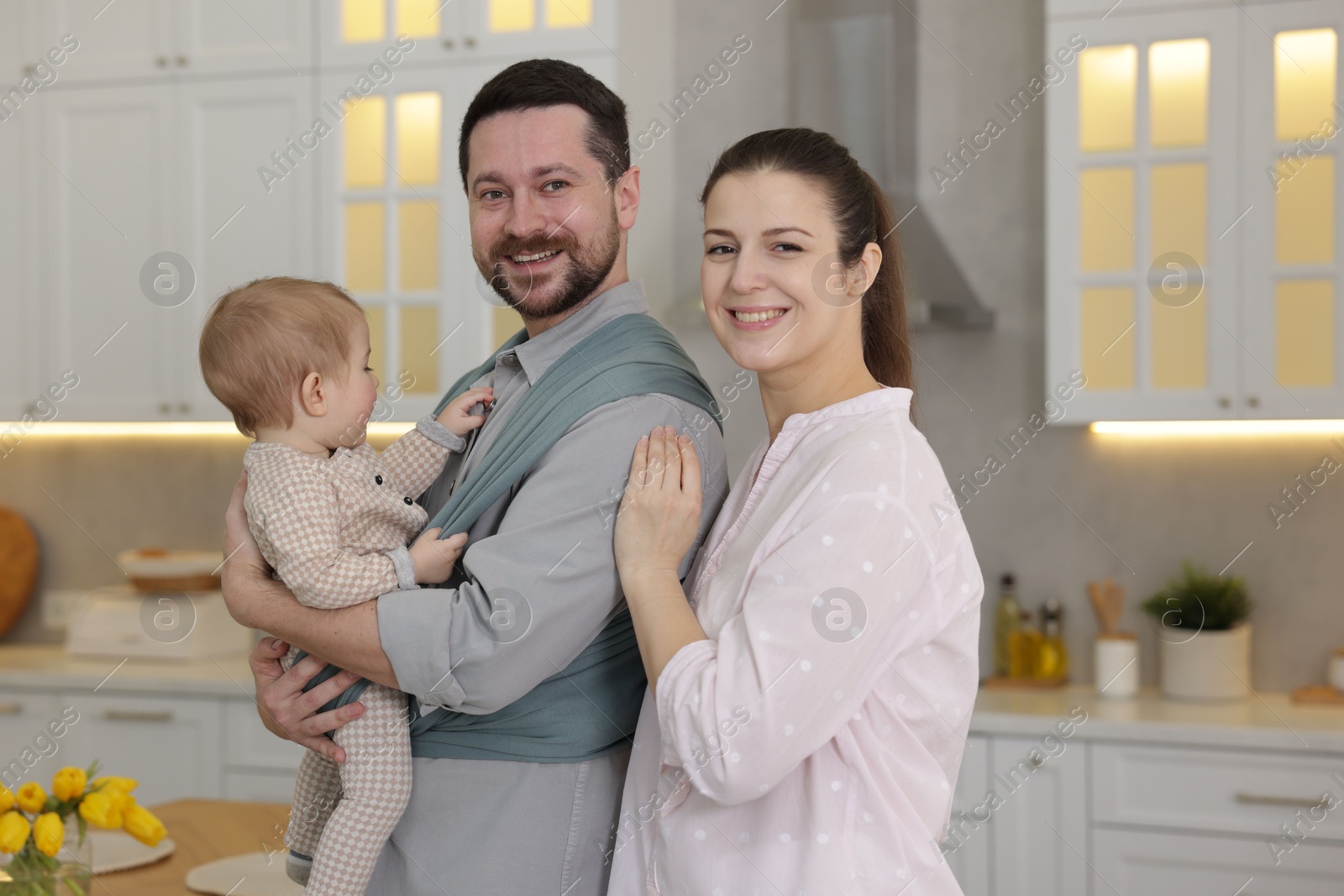 Photo of Portrait of happy family indoors. Father holding his child in sling (baby carrier) at home