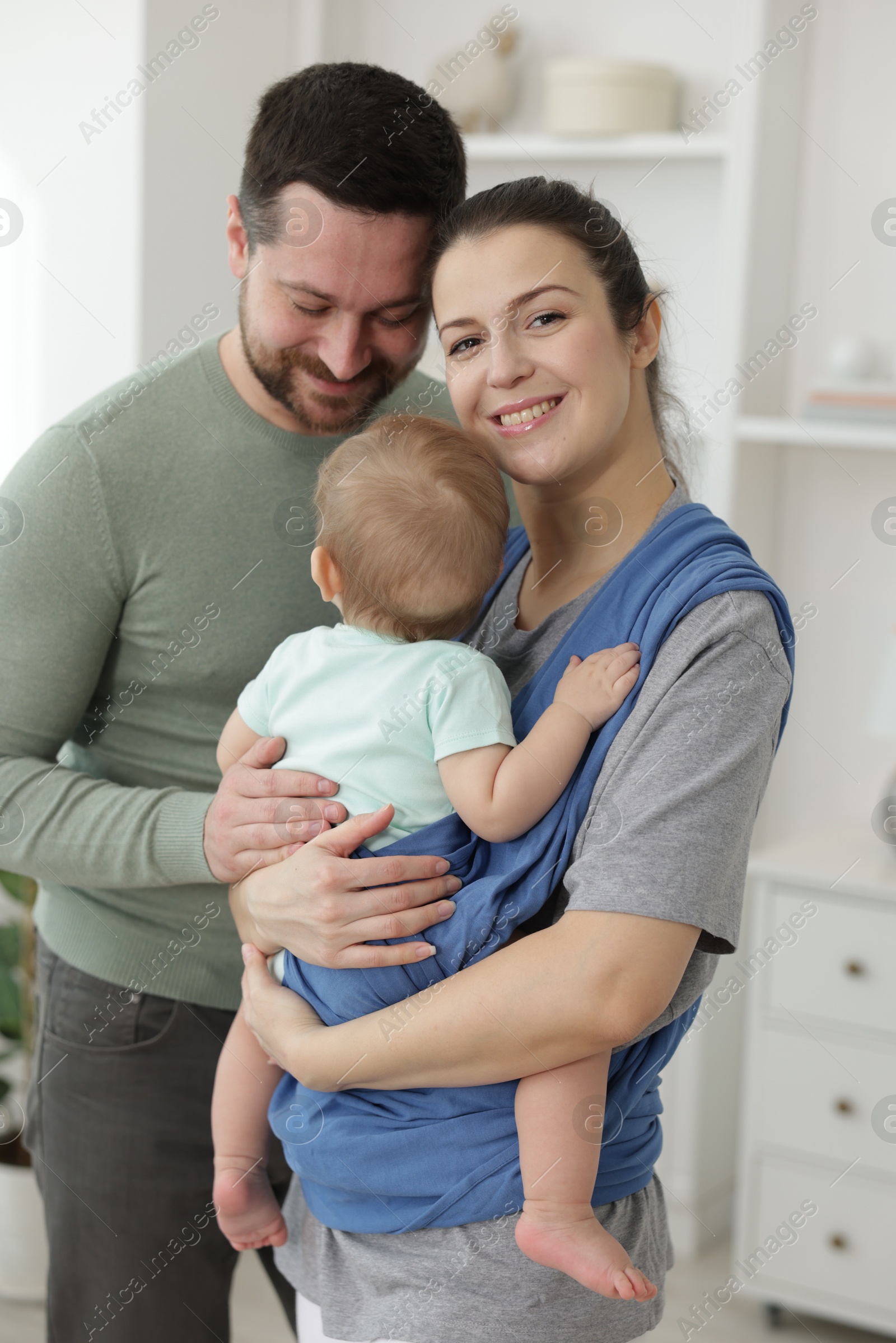 Photo of Portrait of happy family indoors. Mother holding her child in sling (baby carrier) at home