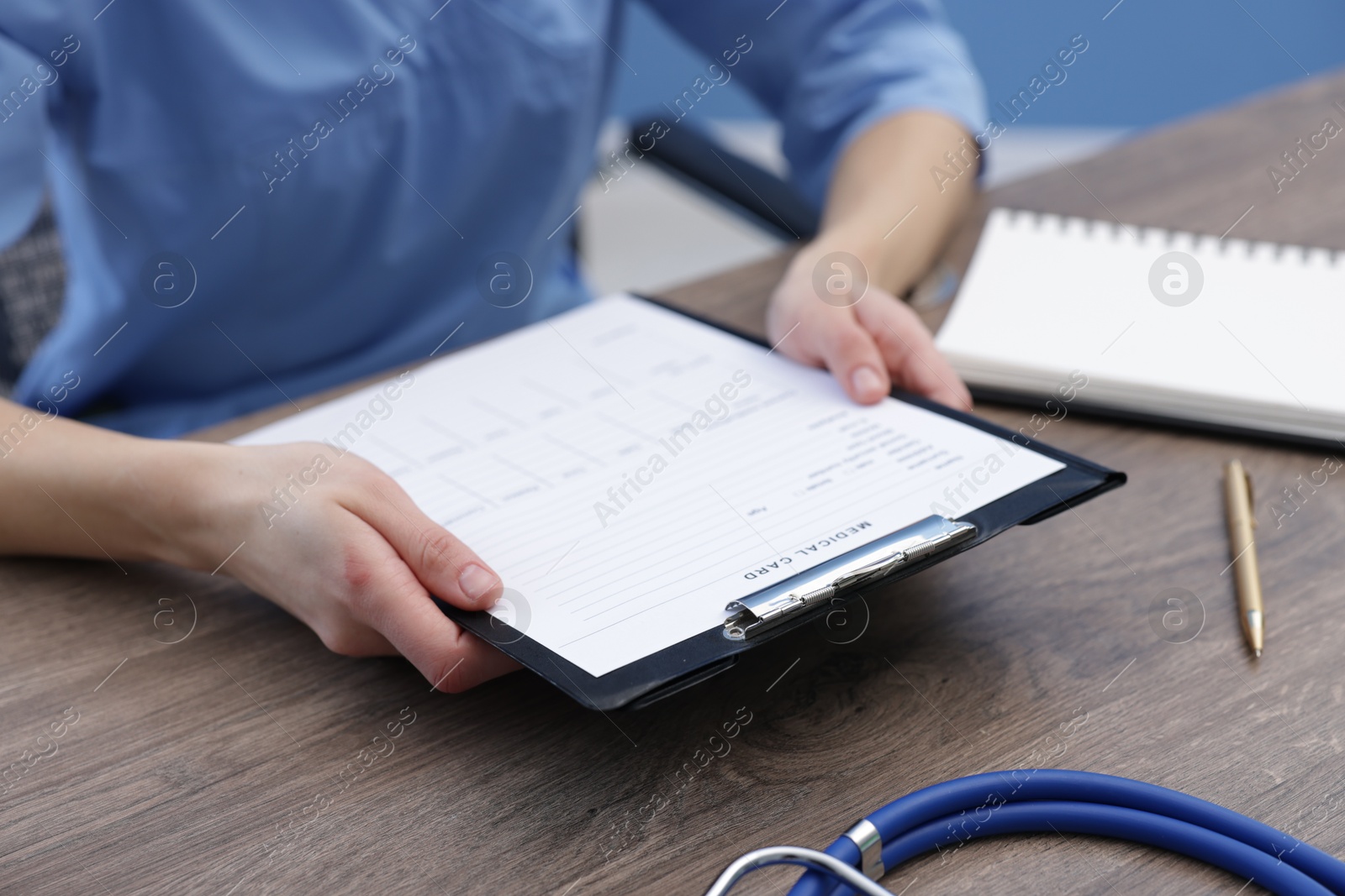 Photo of Doctor holding clipboard with patient's medical card at wooden table in clinic, closeup