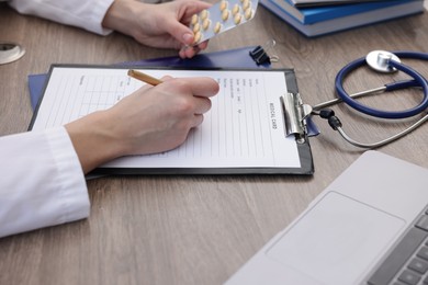 Photo of Doctor with pen and blister of pills filling out patient's medical card at wooden table, closeup