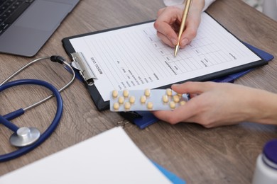 Photo of Doctor with pen and blister of pills filling out patient's medical card at wooden table, closeup